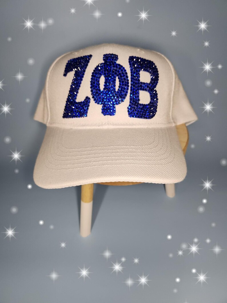 Puede incluir: Gorra de b&eacute;isbol blanca con letras de pedrer&iacute;a azul que dicen "ZФB".