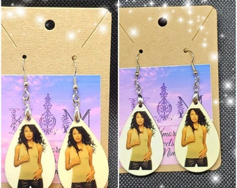 May include: A pair of teardrop-shaped earrings with a white background and a photo of a woman with long dark hair wearing a light brown top and black pants. The earrings are silver and have a hook closure.