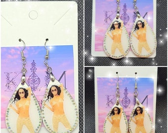 May include: A pair of silver teardrop-shaped earrings with a white background and a photo of a woman in a gold outfit. The earrings are decorated with rhinestones.