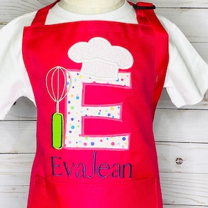 May include: A pink child's apron with a white chef's hat, a green whisk, and a pink letter E with colorful polka dots. The apron has a pocket with the name "EvaJean" embroidered in purple.
