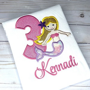 May include: A pink glittery number three with a mermaid with purple and pink scales and blonde hair. The mermaid is holding a purple flower. The name "Kennadi" is embroidered in pink below the number and mermaid.