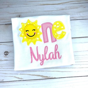 Puede incluir: Un body blanco con un sol amarillo con una cara sonriente y lunares rosas. La palabra "one" est&aacute; bordada en rosa y amarillo con lunares amarillos. El nombre "Nylah" est&aacute; bordado en rosa.