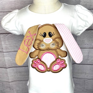 May include: White t-shirt with a brown and pink embroidered bunny with the name "Lex" on the sleeve.