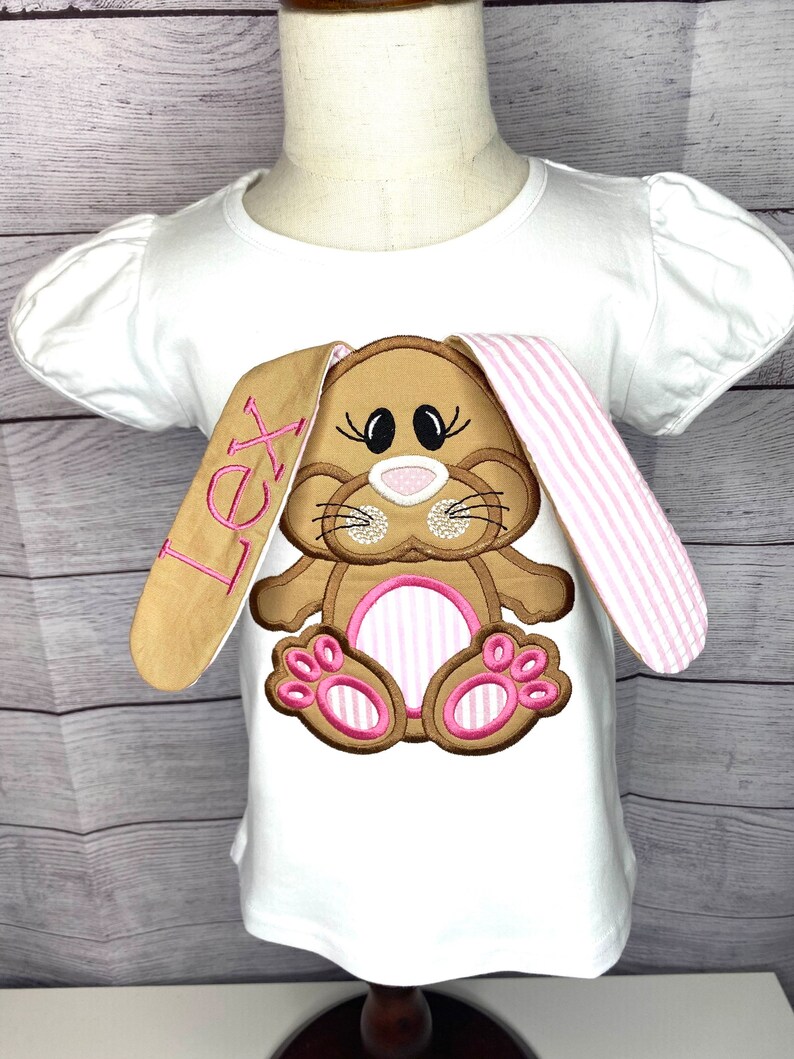 May include: White t-shirt with a brown and pink embroidered bunny with the name "Lex" on the sleeve.