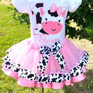 May include: A pink tutu with black and white cow print ribbon trim and a white t-shirt with a black and white cow print number one and a cow face with a pink bow. The t-shirt also has the name "Stormy" embroidered on it.