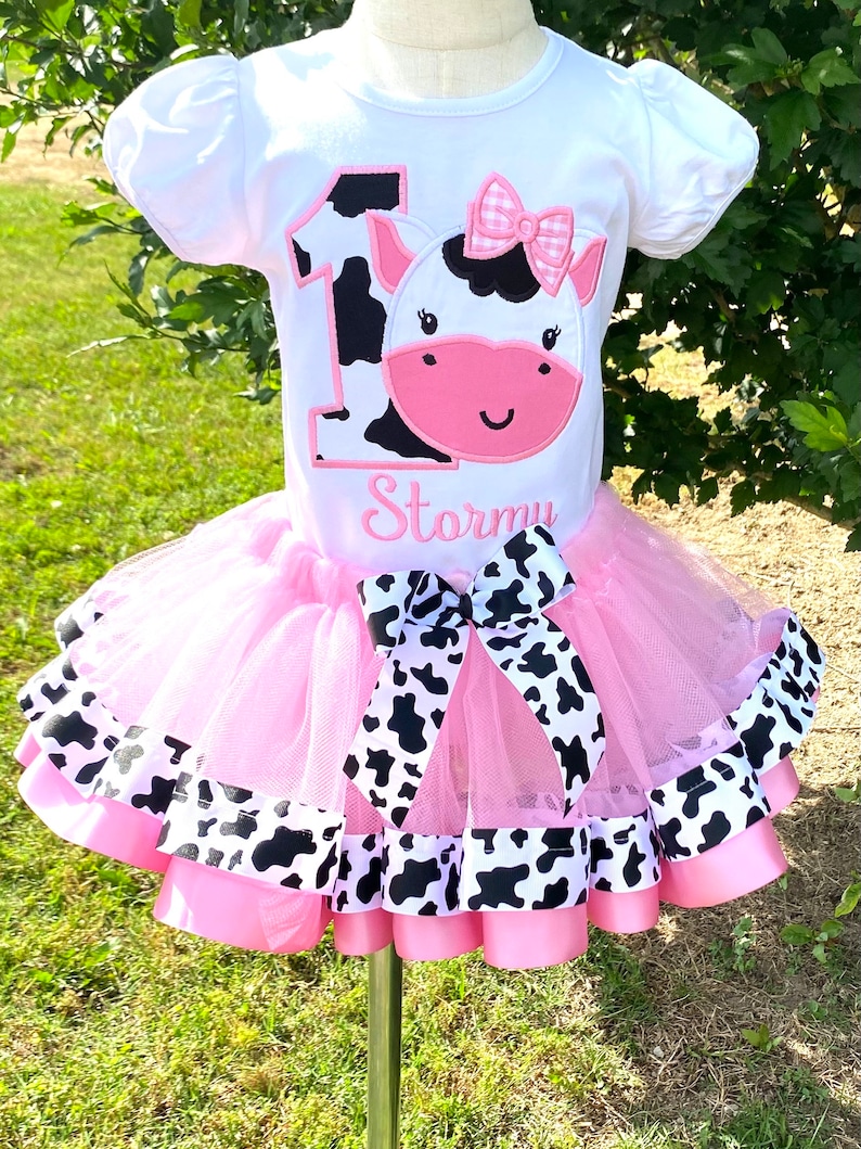 May include: A pink tutu with black and white cow print ribbon trim and a white t-shirt with a black and white cow print number one and a cow face with a pink bow. The t-shirt also has the name "Stormy" embroidered on it.