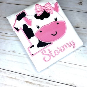 May include: White onesie with a pink cow applique and the name "Stormy" embroidered in pink.