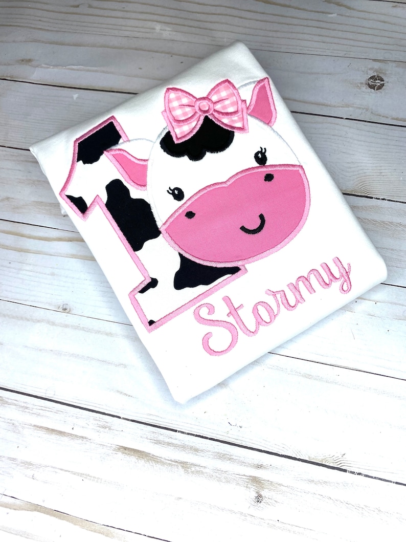 May include: White onesie with a pink cow applique and the name "Stormy" embroidered in pink.