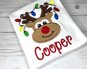 May include: White t-shirt with an embroidered reindeer wearing a string of Christmas lights. The reindeer has a red nose and brown antlers. The name "Cooper" is embroidered in red below the reindeer.