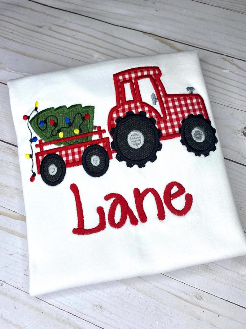 May include: Red and white gingham tractor pulling a wagon with a green Christmas tree and lights. The name "Lane" is embroidered below the tractor.