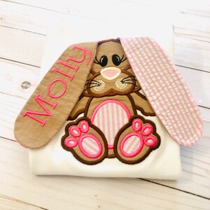 May include: White onesie with a brown and pink embroidered bunny rabbit with pink striped ears. The bunny has a pink nose and pink eyes. The onesie has the name "Molly" embroidered on one of the bunny ears.