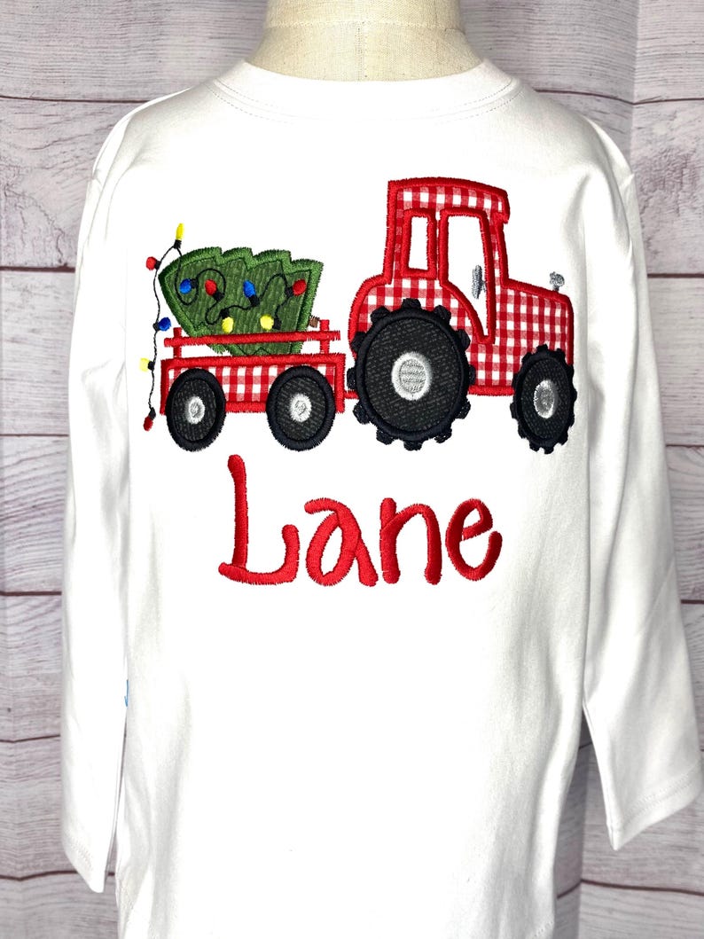 May include: White long-sleeved shirt with a red and white gingham tractor pulling a Christmas tree with lights. The name "Lane" is embroidered in red below the tractor.