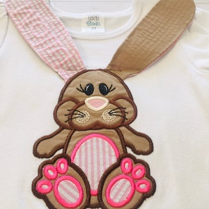 May include: White long-sleeved shirt with a brown and pink embroidered bunny applique. The bunny has pink and white striped ears, a pink nose, and pink and white striped feet.