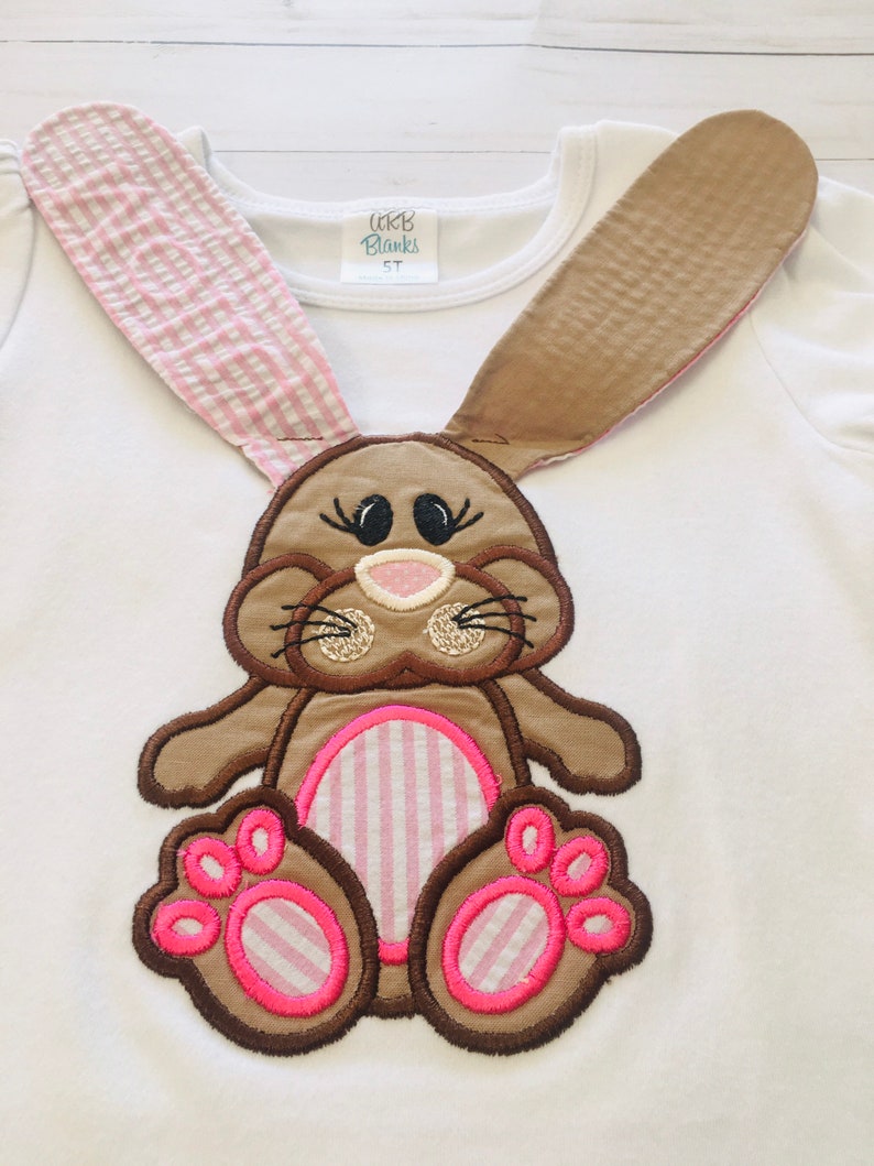 May include: White long-sleeved shirt with a brown and pink embroidered bunny applique. The bunny has pink and white striped ears, a pink nose, and pink and white striped feet.