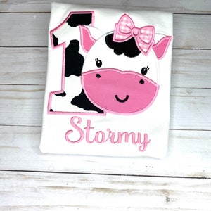 May include: A white onesie with a pink cow applique. The cow has black spots and a pink bow. The onesie is personalized with the name "Stormy" in pink.