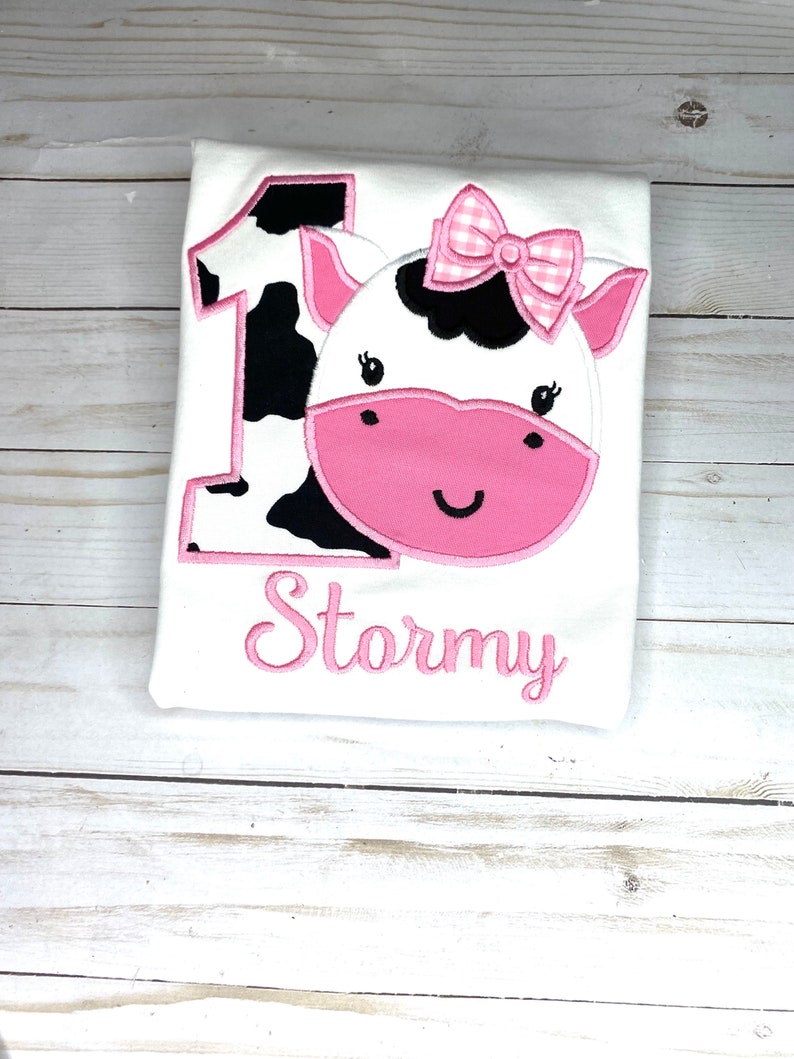 May include: A white onesie with a pink cow applique. The cow has black spots and a pink bow. The onesie is personalized with the name "Stormy" in pink.