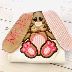 May include: A white t-shirt with a brown bunny rabbit embroidered on the front. The bunny has pink and white striped ears and paws. The name "Molly" is embroidered on one of the bunny's ears.