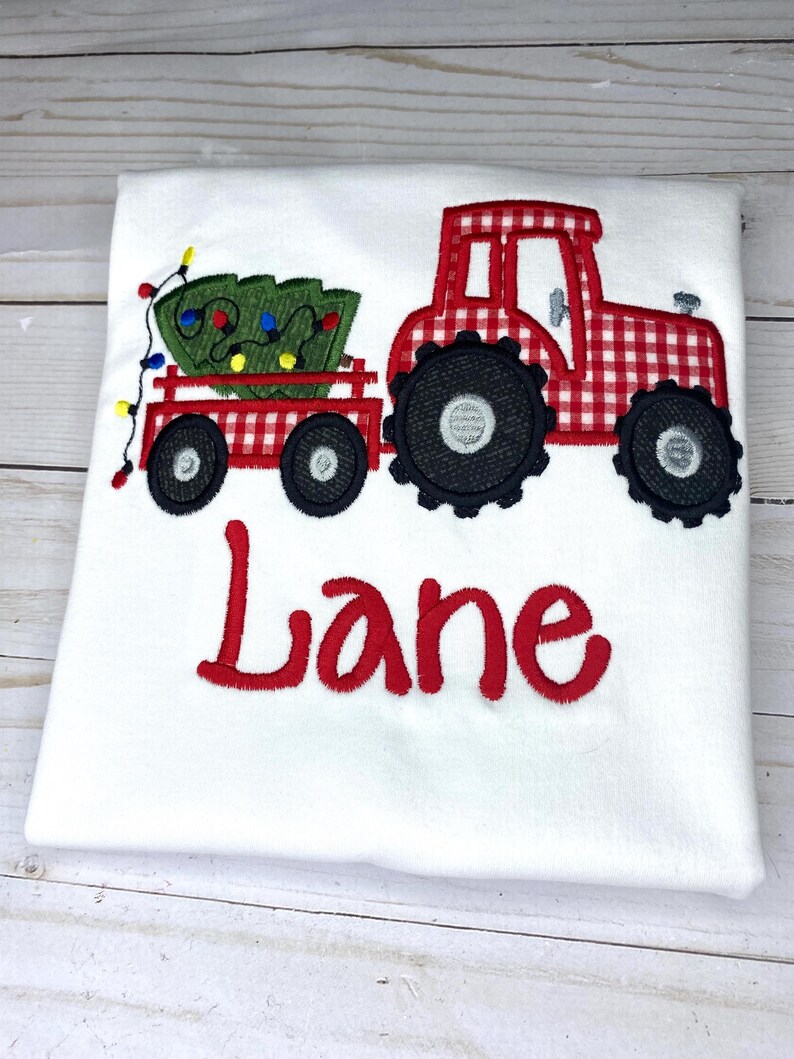 May include: Red and white gingham tractor pulling a wagon with a green Christmas tree and lights. The name "Lane" is embroidered below the tractor.