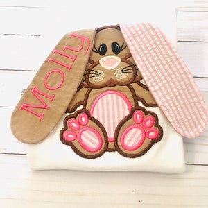 May include: A white onesie with a brown and pink embroidered bunny with pink and white striped ears. The bunny's ears are long and have the name "Molly" embroidered on one ear in pink thread.