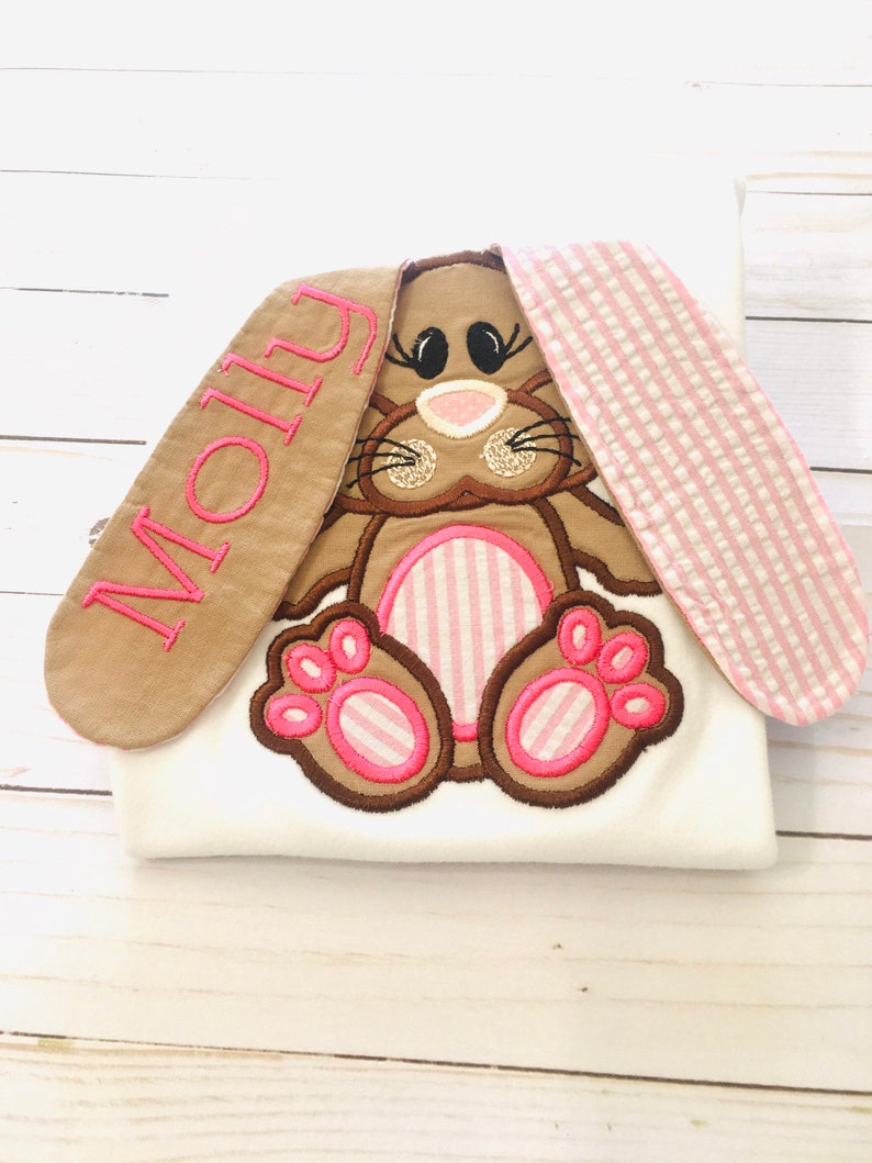May include: A white onesie with a brown and pink embroidered bunny with pink and white striped ears. The bunny's ears are long and have the name "Molly" embroidered on one ear in pink thread.