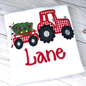 May include: Red and white gingham tractor pulling a wagon with a green Christmas tree and colorful lights. The name "Lane" is embroidered below the tractor.
