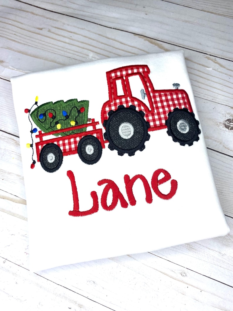 May include: Red and white gingham tractor pulling a wagon with a green Christmas tree and colorful lights. The name "Lane" is embroidered below the tractor.