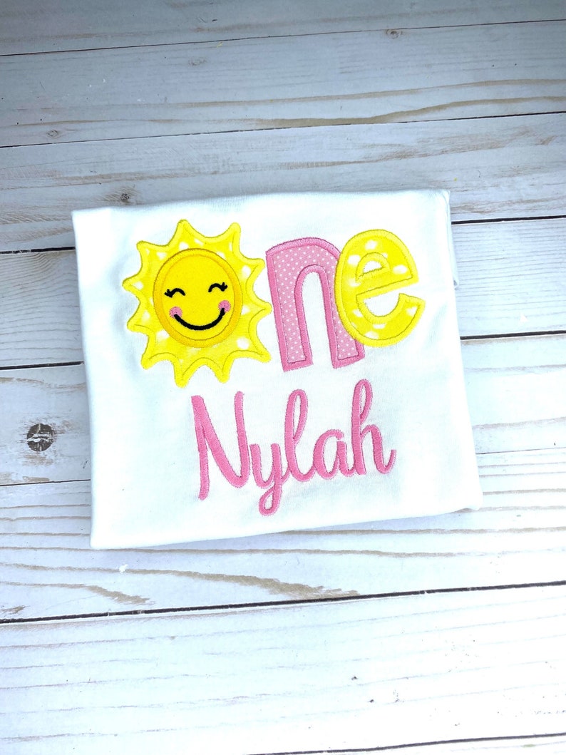 Puede incluir: Un body blanco con un sol amarillo con una cara sonriente y lunares rosas. La palabra "one" est&aacute; bordada en rosa y amarillo con lunares amarillos. El nombre "Nylah" est&aacute; bordado en rosa.
