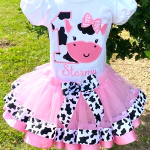 May include: A pink tutu with black and white cow print ribbon trim. A white t-shirt with a black and white cow print number one and a pink cow face with a pink bow. The shirt says "Stormy" underneath the cow.