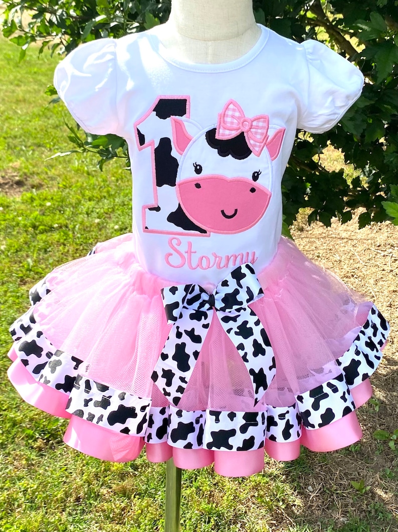 May include: A pink tutu with black and white cow print ribbon trim. A white t-shirt with a black and white cow print number one and a pink cow face with a pink bow. The shirt says "Stormy" underneath the cow.