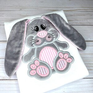 May include: A white t-shirt with a gray and pink embroidered bunny rabbit design. The bunny has long ears, pink stripes on its body, and pink paw prints.