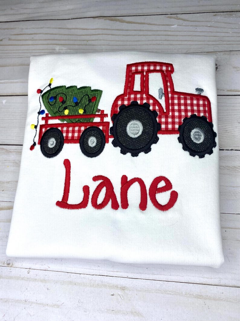 May include: Red and white gingham tractor pulling a wagon with a green Christmas tree and lights. The name "Lane" is embroidered below the tractor.