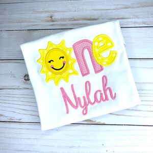 Puede incluir: Un body blanco con un sol bordado en rosa y amarillo y la palabra "one" en rosa con lunares blancos. El nombre "Nylah" est&aacute; bordado en rosa debajo de la palabra "one".