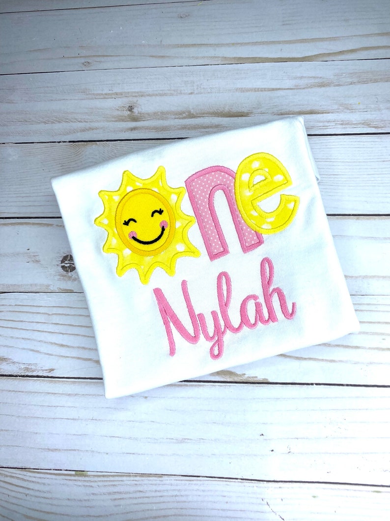 Puede incluir: Un body blanco con un sol bordado en rosa y amarillo y la palabra "one" en rosa con lunares blancos. El nombre "Nylah" est&aacute; bordado en rosa debajo de la palabra "one".