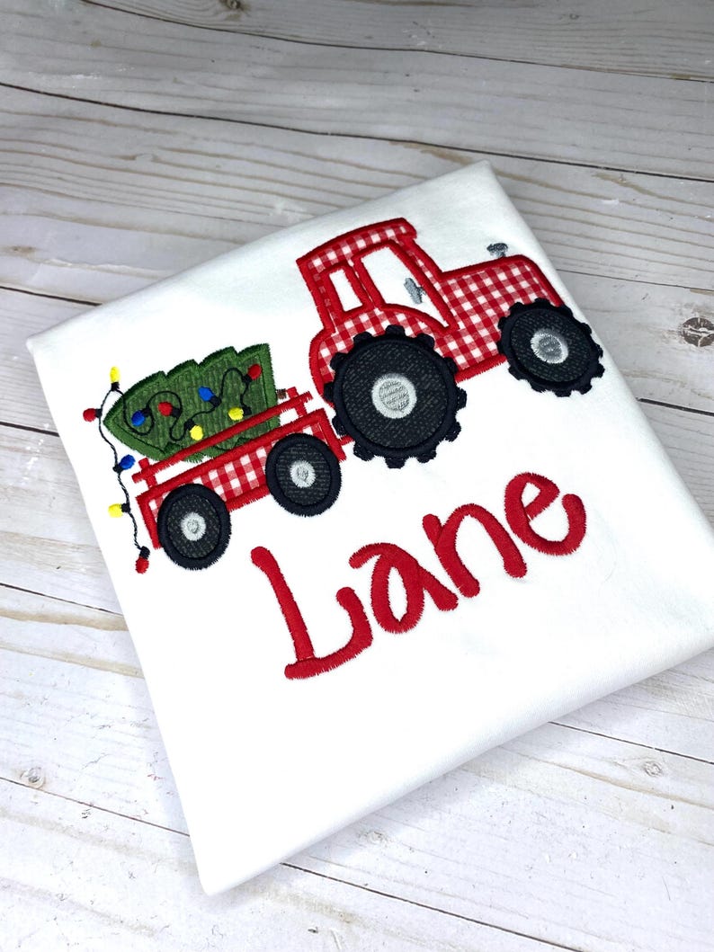 May include: Red and white gingham tractor pulling a wagon with a green Christmas tree and lights. The name "Lane" is embroidered in red below the tractor.
