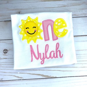 Puede incluir: Un body blanco con un sol amarillo con una cara sonriente, la palabra "one" en rosa con lunares blancos y el nombre "Nylah" en rosa.