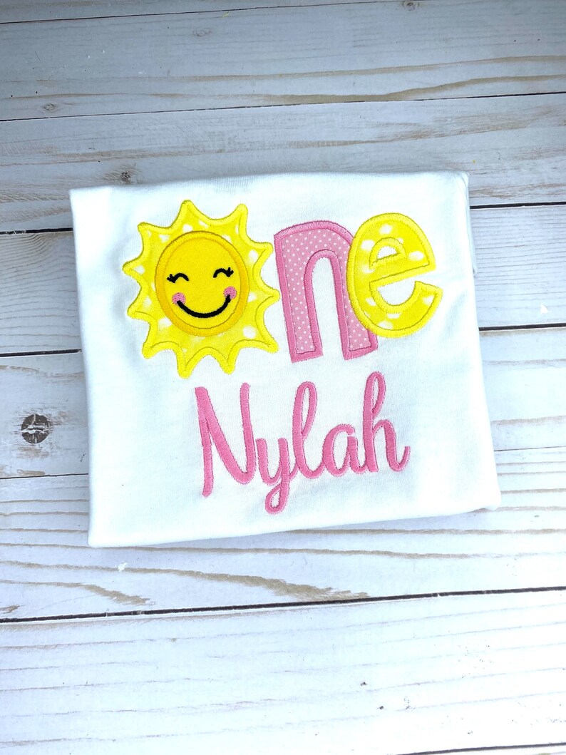 Puede incluir: Un body blanco con un sol amarillo con una cara sonriente, la palabra "one" en rosa con lunares blancos y el nombre "Nylah" en rosa.