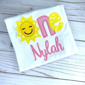 Puede incluir: Un body blanco con un sol bordado en rosa y amarillo, la palabra "one" en rosa con lunares blancos y el nombre "Nylah" en rosa.