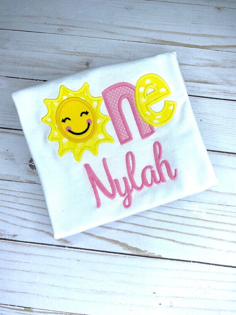 Puede incluir: Un body blanco con un sol bordado en rosa y amarillo, la palabra "one" en rosa con lunares blancos y el nombre "Nylah" en rosa.