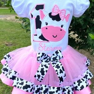 May include: A pink tutu with black and white cow print ribbon trim. A white t-shirt with a black and white cow print number one and a pink cow face with a pink bow. The shirt is personalized with the name "Stormi".