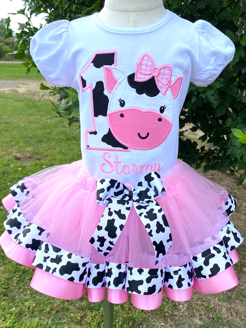 May include: A pink tutu with black and white cow print ribbon trim. A white t-shirt with a black and white cow print number one and a pink cow face with a pink bow. The shirt is personalized with the name "Stormi".
