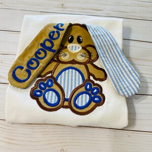 May include: White onesie with a brown bunny embroidered on the front. The bunny has blue and white striped ears and paws. The onesie has the name "Cooper" embroidered on one of the bunny ears.