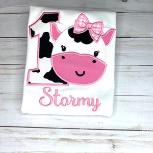 May include: White onesie with a pink cow applique and the number 1. The cow has black and white spots and a pink bow. The onesie is personalized with the name "Stormy" in pink.
