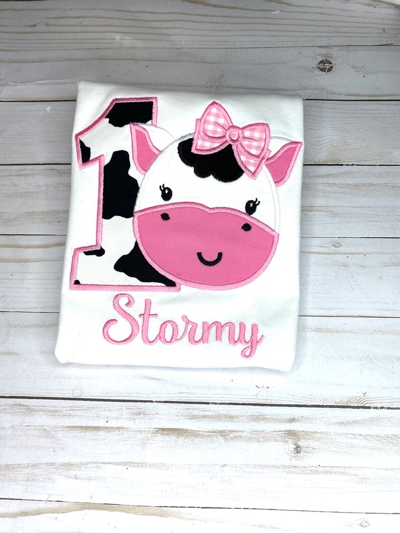 May include: White onesie with a pink cow applique and the number 1. The cow has black and white spots and a pink bow. The onesie is personalized with the name "Stormy" in pink.
