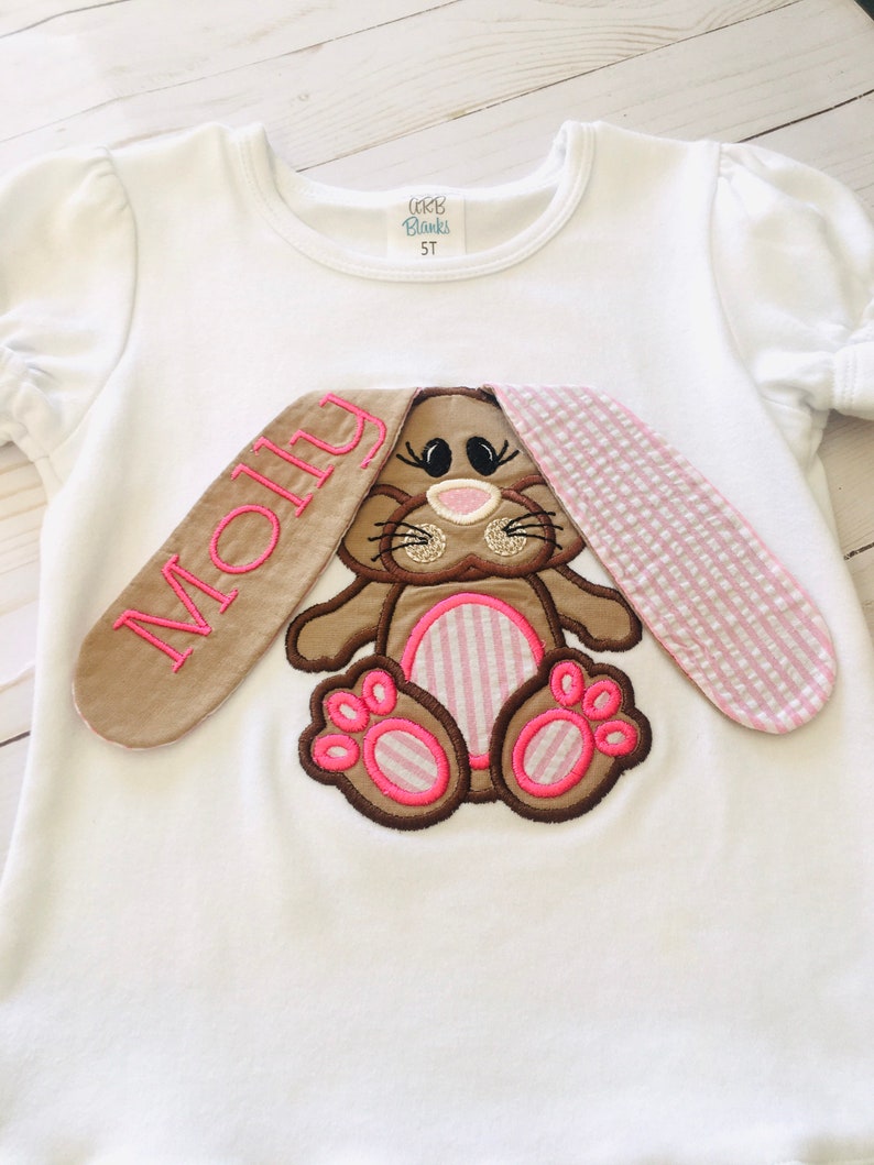 May include: White t-shirt with a brown bunny applique. The bunny has pink ears, pink paws, and a pink nose. The shirt is personalized with the name "Molly" in brown embroidery.