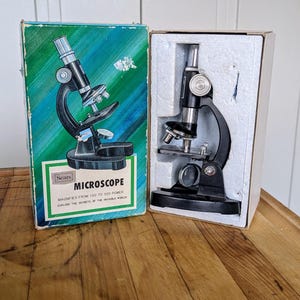 May include: A black microscope in its original box. The box is green and blue with the words "Sears Microscope" and "Magnifies from 100 to 300 power" printed on it. The microscope is a vintage toy.