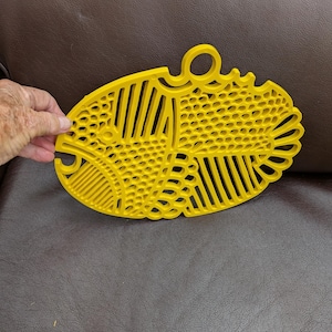 Peut inclure: Un dessous de plat ovale jaune vif arborant un motif de poisson stylisé. La surface est composée de motifs géométriques en relief complexes, incluant des lignes parallèles et des textures en nid d'abeille, avec une boucle circulaire au sommet.