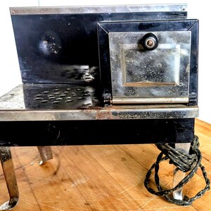 Könnte beinhalten: Ein antiker schwarzer und silberner Metallofen mit einer kleinen Ofentür und einer Reihe von Löchern auf der Oberseite. Der Ofen hat vier gebogene Beine und ein Netzkabel, das um ein Bein gewickelt ist. Der Ofen steht auf einer Holzoberfläche.
