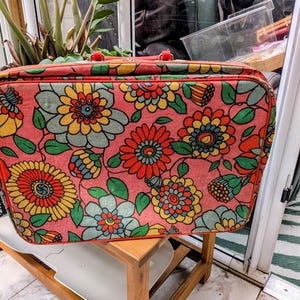 May include: A rectangular suitcase featuring a vibrant floral pattern with red, yellow, and light blue flowers on a pink background. The luggage has rounded corners, a red fabric trim along the edges, and a red handle attached to the top.