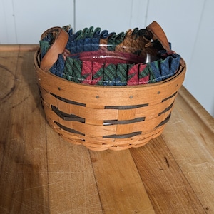 Puede incluir: Una cesta redonda tejida con exterior de madera marrón y forro de tela estampada. La cesta tiene dos asas de cuero y un borde con volantes. El forro de tela presenta un estampado rojo, verde y azul. La cesta está sobre una superficie de madera.