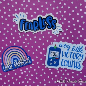 May include: Three blue and white vinyl stickers with inspirational messages. The first sticker says "Fearless" in a blue and white font. The second sticker says "Chase Rainbows" with a rainbow graphic. The third sticker says "Every little victory counts" with a blue and white graphic of a blood glucose meter.